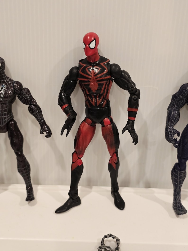 May include: A red and black Spider-Man action figure with a spider emblem on the chest. The figure has articulated joints and is posed with arms extended. Other Spider-Man figures are visible in the background.