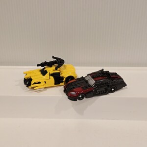 May include: Two toy vehicles: a yellow armored truck with black details and a black and red sports car. Both toys have wheels and are on a white surface. The yellow truck has a weapon on top.