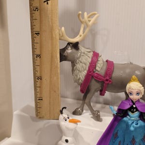 May include: A collection of Frozen-themed figurines, including a reindeer with a pink harness, a snowman, and a doll of Elsa in a blue dress and purple cape. A wooden ruler is placed next to the reindeer.