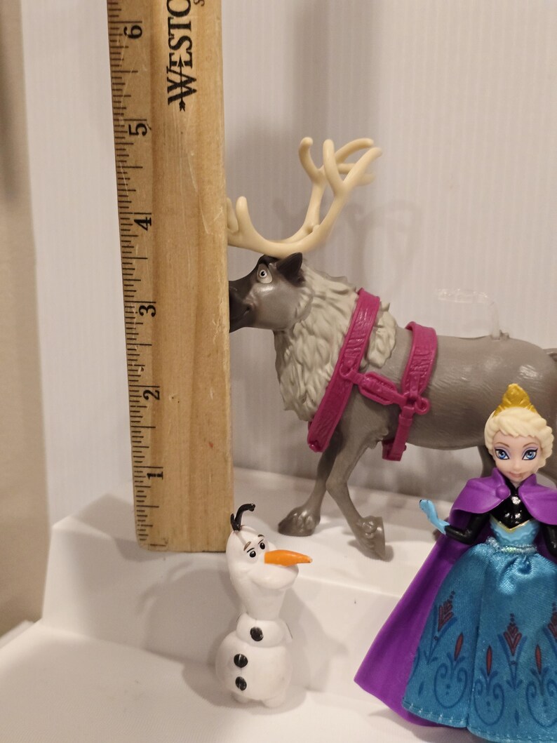 May include: A collection of Frozen-themed figurines, including a reindeer with a pink harness, a snowman, and a doll of Elsa in a blue dress and purple cape. A wooden ruler is placed next to the reindeer.