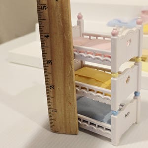 Puede incluir: Una cama de mu&ntilde;ecas blanca de tres niveles con ropa de cama rosa, amarilla y azul. La cama mide aproximadamente 13 cm de alto. Cada cama tiene un recorte en forma de coraz&oacute;n y est&aacute; decorada con detalles pastel.