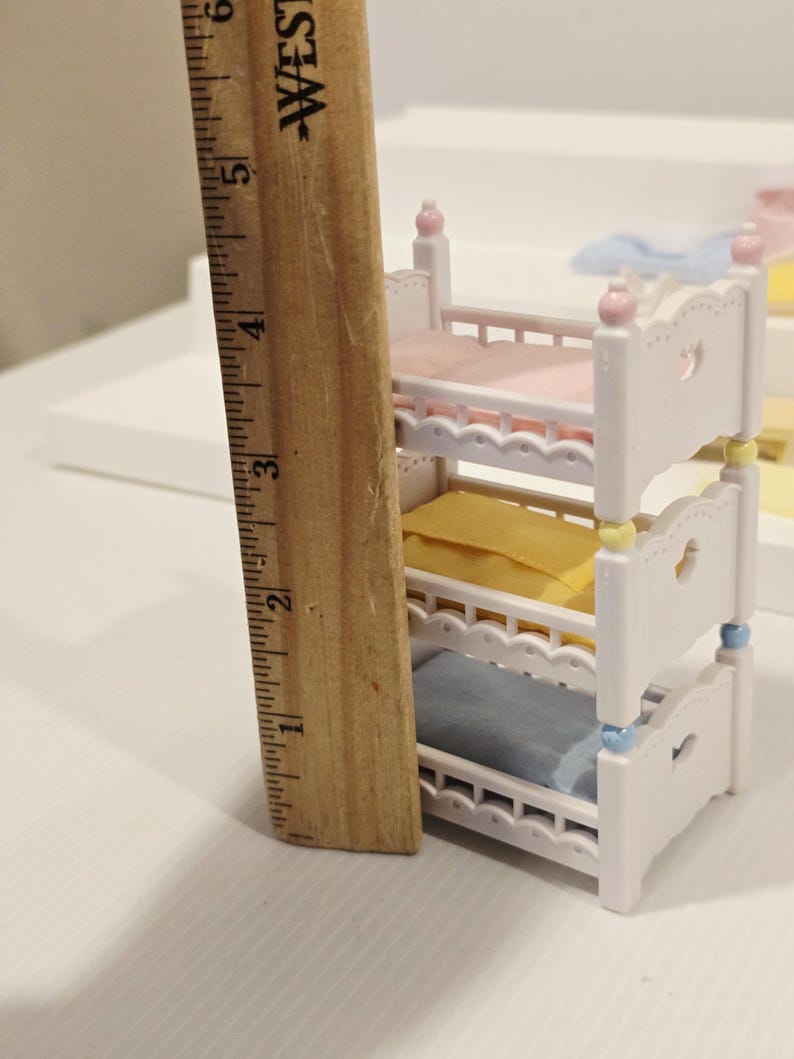 Puede incluir: Una cama de mu&ntilde;ecas blanca de tres niveles con ropa de cama rosa, amarilla y azul. La cama mide aproximadamente 13 cm de alto. Cada cama tiene un recorte en forma de coraz&oacute;n y est&aacute; decorada con detalles pastel.