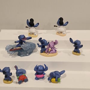 May include: A collection of blue Stitch figurines in various poses. Some are surfing, wearing leis, or holding objects. The figurines are on small, round bases, and some are paired with other characters. The overall theme is tropical and playful.