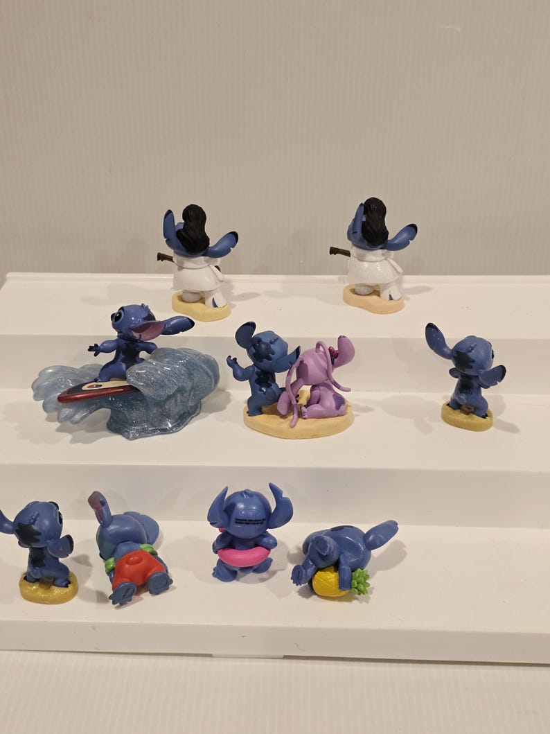 May include: A collection of blue Stitch figurines in various poses. Some are surfing, wearing leis, or holding objects. The figurines are on small, round bases, and some are paired with other characters. The overall theme is tropical and playful.