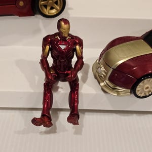 May include: A red and gold Iron Man action figure sits on a white surface. The figure is in a seated position, with its legs dangling. A red car with gold accents is visible in the background.