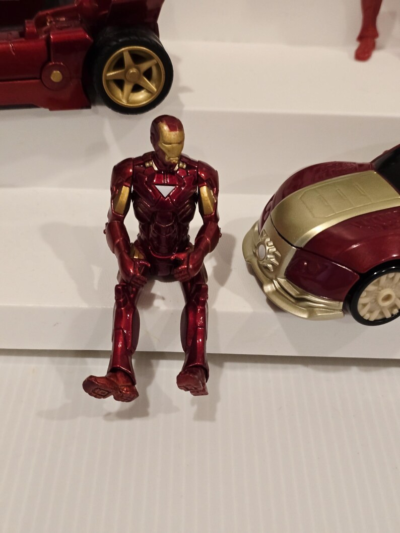 May include: A red and gold Iron Man action figure sits on a white surface. The figure is in a seated position, with its legs dangling. A red car with gold accents is visible in the background.