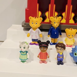 K&ouml;nnte beinhalten: Eine Sammlung von Daniel Tiger's Nachbarschaftsfiguren, darunter Daniel Tiger, Katerina Kittycat und O die Eule. Die Figuren sind farbenfroh und stehen vor einem roten Eisenbahnwagen.