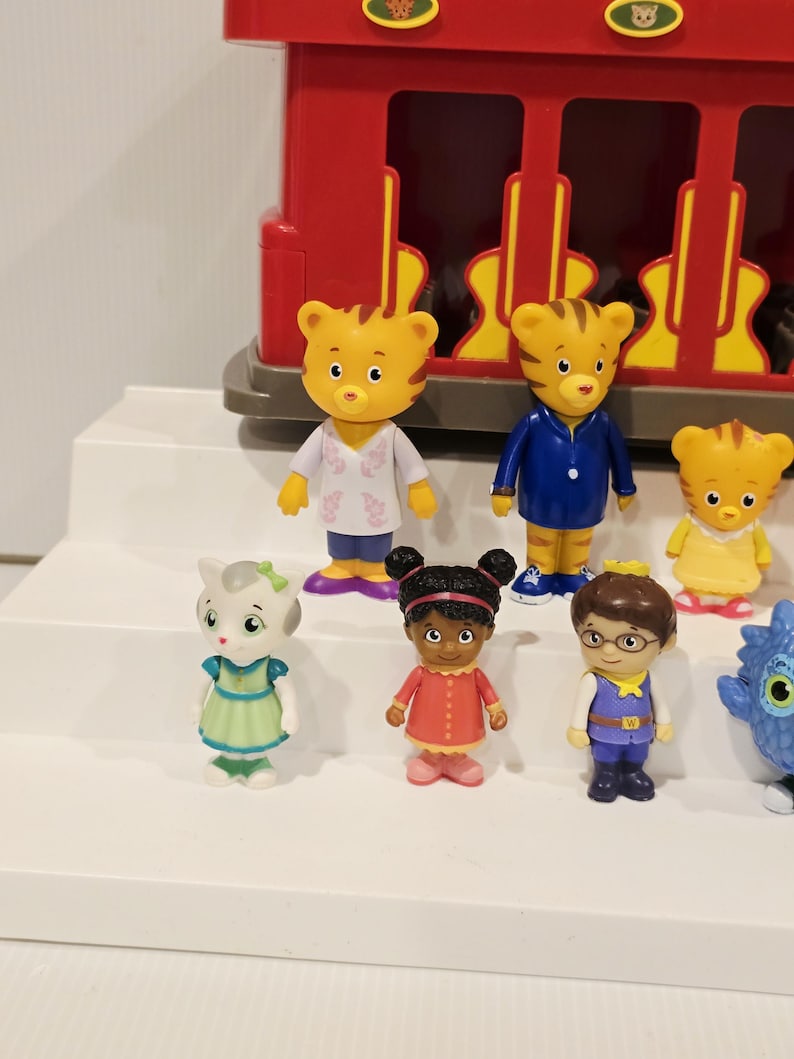 K&ouml;nnte beinhalten: Eine Sammlung von Daniel Tiger's Nachbarschaftsfiguren, darunter Daniel Tiger, Katerina Kittycat und O die Eule. Die Figuren sind farbenfroh und stehen vor einem roten Eisenbahnwagen.