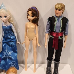 May include: A selection of dolls based on characters from the animated film "Frozen." The dolls include Elsa in a blue dress, Anna, a doll with brown hair and a purple headband, and Kristoff in a black and blue outfit.
