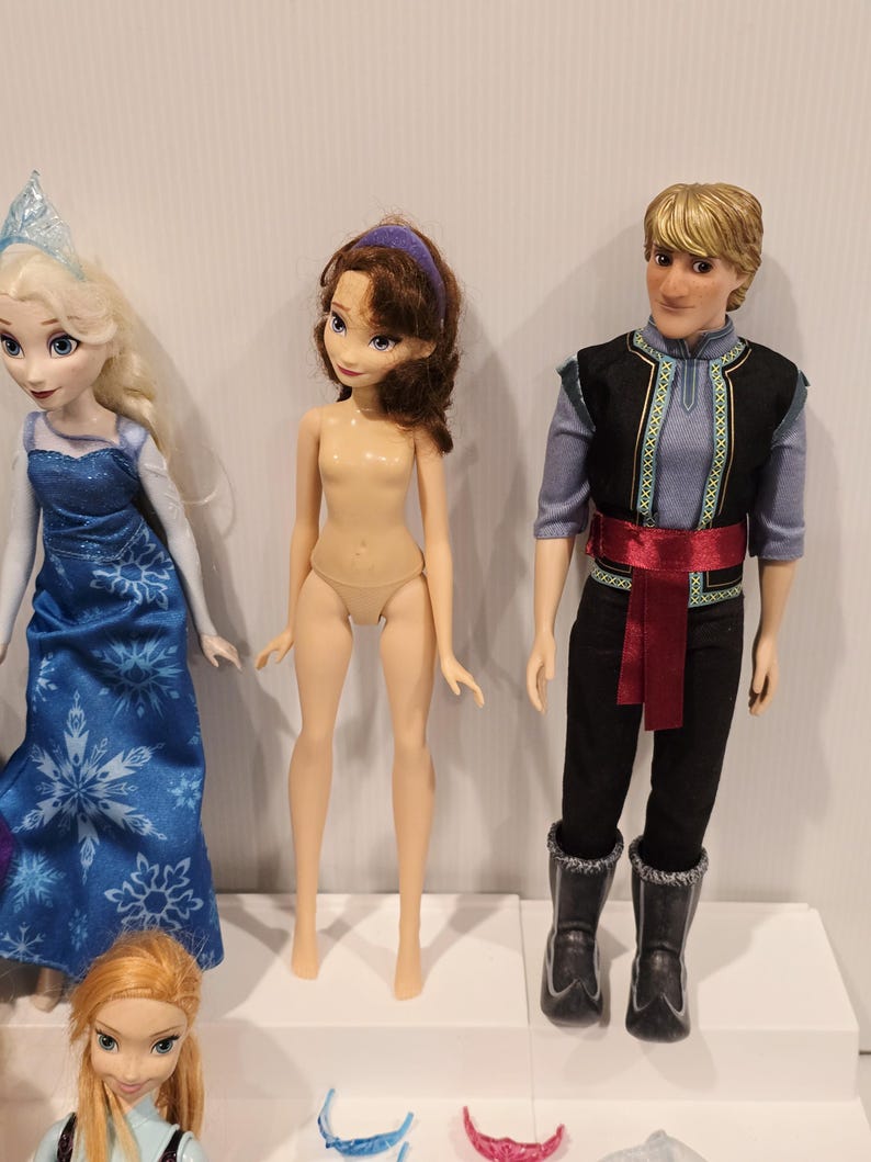 May include: A selection of dolls based on characters from the animated film "Frozen." The dolls include Elsa in a blue dress, Anna, a doll with brown hair and a purple headband, and Kristoff in a black and blue outfit.