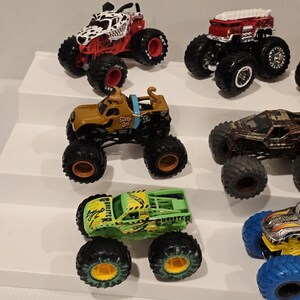 Op de afbeelding: Een verzameling speelgoed monstertrucks in verschillende kleuren, waaronder rood, bruin, groen en zwart. De trucks hebben grote banden en gedetailleerde ontwerpen, zoals een truck met Dalmatische thema en een Scooby-Doo truck.