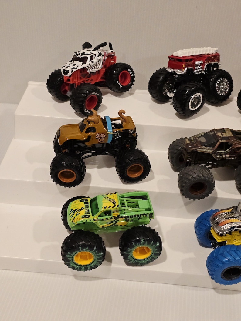 Op de afbeelding: Een verzameling speelgoed monstertrucks in verschillende kleuren, waaronder rood, bruin, groen en zwart. De trucks hebben grote banden en gedetailleerde ontwerpen, zoals een truck met Dalmatische thema en een Scooby-Doo truck.