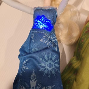 May include: A doll in a blue dress with snowflake patterns and a glowing blue centre. The doll has blonde hair and is wearing a blue dress with white snowflake patterns. The doll's arms are white.