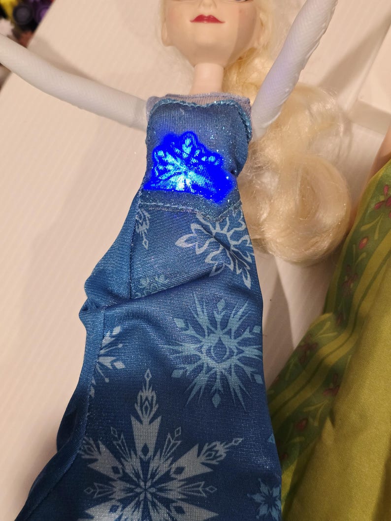 May include: A doll in a blue dress with snowflake patterns and a glowing blue centre. The doll has blonde hair and is wearing a blue dress with white snowflake patterns. The doll's arms are white.
