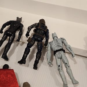 May include: Three action figures: a black superhero, a figure in dark blue tactical gear, and a gray superhero with a bat-like cowl. The figures are posed on a white surface, suggesting a toy or collectible theme.