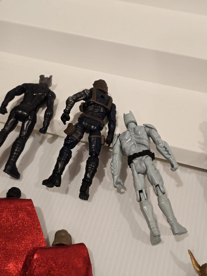 May include: Three action figures: a black superhero, a figure in dark blue tactical gear, and a gray superhero with a bat-like cowl. The figures are posed on a white surface, suggesting a toy or collectible theme.
