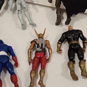 May include: A collection of superhero action figures. The figures include Captain America in red, white, and blue, a figure in red and gold, and a figure in black and gold. Other figures are also visible.