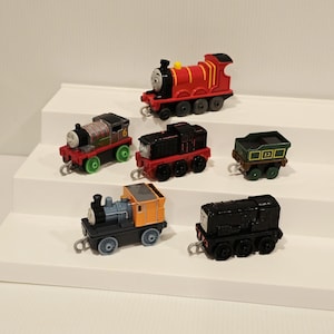 May include: A collection of six small toy trains and a cargo car. The set includes a red engine, a grey and red engine with the number 6, a black engine, a green cargo car with the number 12, an orange and blue engine, and a black engine.