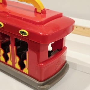 K&ouml;nnte beinhalten: Ein roter Spielzeug-Trolleybus mit gelbem Griff. Der Trolleybus hat gelbe und schwarze Akzente und eine graue Basis. Das Design umfasst offene Seiten und eine abgerundete Front.