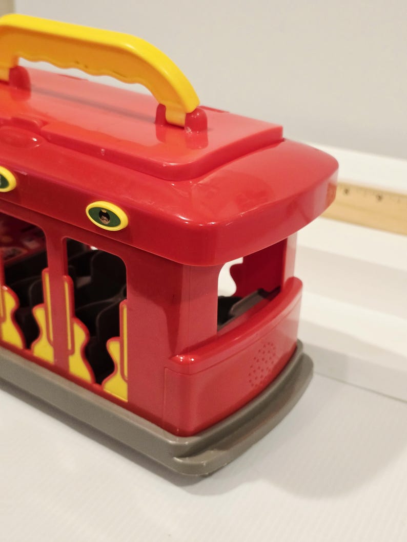 K&ouml;nnte beinhalten: Ein roter Spielzeug-Trolleybus mit gelbem Griff. Der Trolleybus hat gelbe und schwarze Akzente und eine graue Basis. Das Design umfasst offene Seiten und eine abgerundete Front.