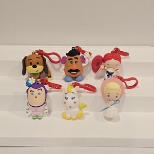 May include: A selection of six character keyrings. The keyrings feature characters from the Toy Story franchise, including a dog, Mr. Potato Head, Jessie, Buzz Lightyear, a unicorn, and Bo Peep. Each keyring has a red loop.