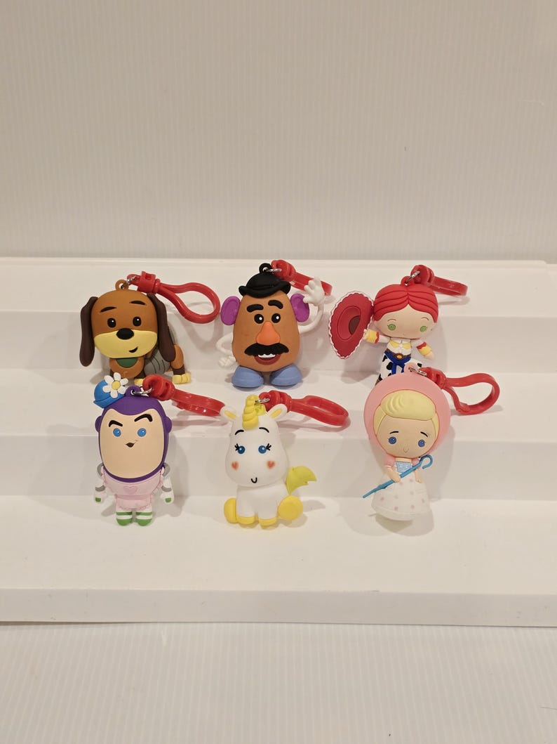 May include: A selection of six character keyrings. The keyrings feature characters from the Toy Story franchise, including a dog, Mr. Potato Head, Jessie, Buzz Lightyear, a unicorn, and Bo Peep. Each keyring has a red loop.