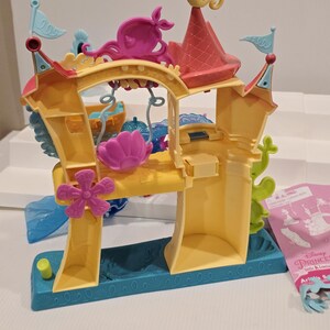 K&ouml;nnte beinhalten: Ein gelbes und t&uuml;rkisfarbenes Kunststoff-Spielset mit Burgdesign. Das Spielset verf&uuml;gt &uuml;ber rosa und lila dekorative Elemente, darunter ein Seepferdchen, einen Oktopus und eine Blume. Kleine blaue Fahnen und ein rotes Dach vervollst&auml;ndigen das Design. Teil der Disney Princess Little Kingdom-Serie.