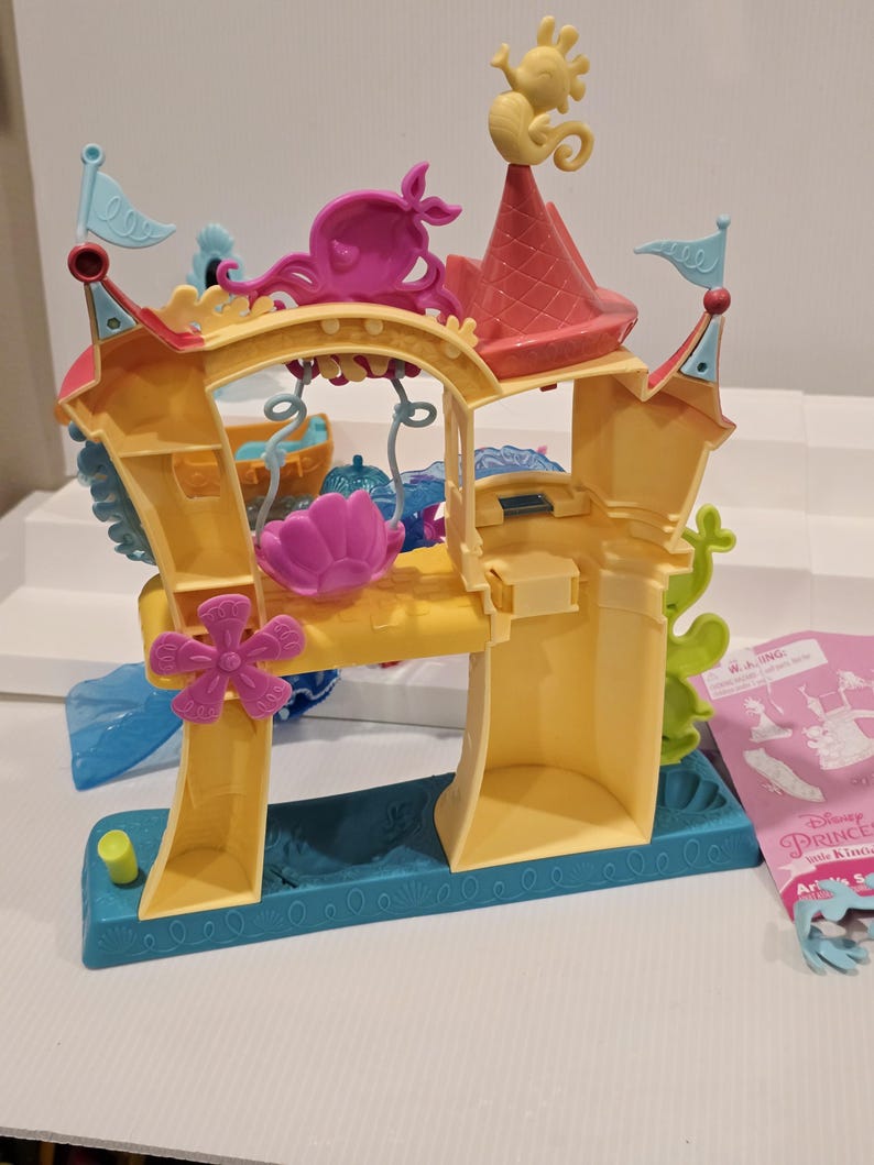 K&ouml;nnte beinhalten: Ein gelbes und t&uuml;rkisfarbenes Kunststoff-Spielset mit Burgdesign. Das Spielset verf&uuml;gt &uuml;ber rosa und lila dekorative Elemente, darunter ein Seepferdchen, einen Oktopus und eine Blume. Kleine blaue Fahnen und ein rotes Dach vervollst&auml;ndigen das Design. Teil der Disney Princess Little Kingdom-Serie.
