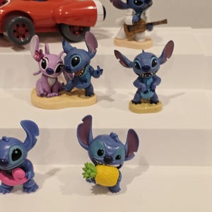 May include: A collection of Stitch figurines in various poses. One figurine is in a red race car, another is dressed as Elvis Presley, and others are holding objects like a pineapple and a pink donut. The figurines are on a white surface.