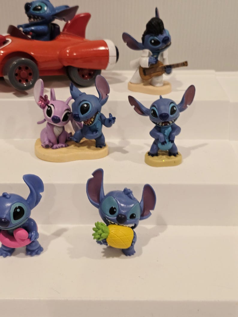 May include: A collection of Stitch figurines in various poses. One figurine is in a red race car, another is dressed as Elvis Presley, and others are holding objects like a pineapple and a pink donut. The figurines are on a white surface.