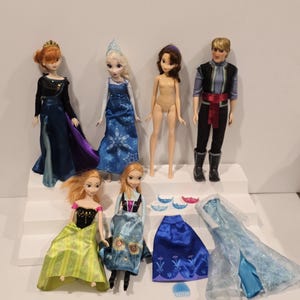 May include: A selection of Disney Frozen dolls and accessories. The dolls feature Elsa, Anna, Kristoff, and other characters. Accessories include dresses, crowns, and other items. The dolls are displayed on a white tiered platform.