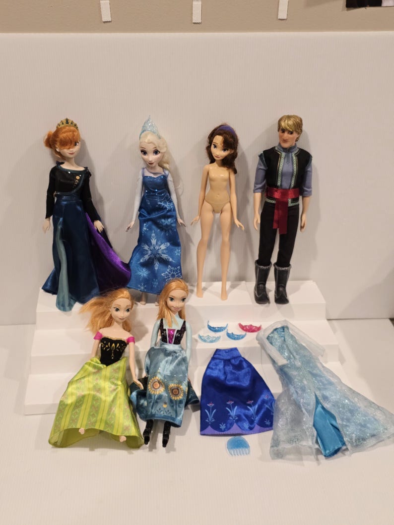 May include: A selection of Disney Frozen dolls and accessories. The dolls feature Elsa, Anna, Kristoff, and other characters. Accessories include dresses, crowns, and other items. The dolls are displayed on a white tiered platform.