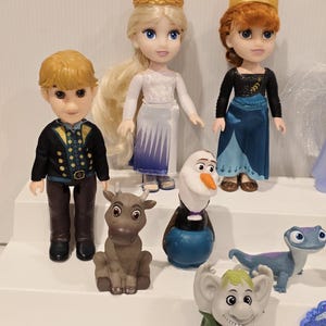 May include: A collection of Disney Frozen character figurines, including Elsa, Anna, Kristoff, Olaf, Sven, Bruni, and a troll. Elsa and Anna wear their signature dresses and golden crowns. The figurines are on a white surface.