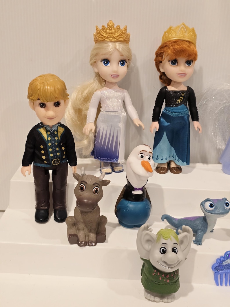 May include: A collection of Disney Frozen character figurines, including Elsa, Anna, Kristoff, Olaf, Sven, Bruni, and a troll. Elsa and Anna wear their signature dresses and golden crowns. The figurines are on a white surface.