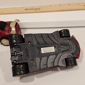 May include: A red toy car with black wheels and a gold spoiler. The underside of the car is visible, showing a textured design and a warning label. A wooden ruler is in the background.
