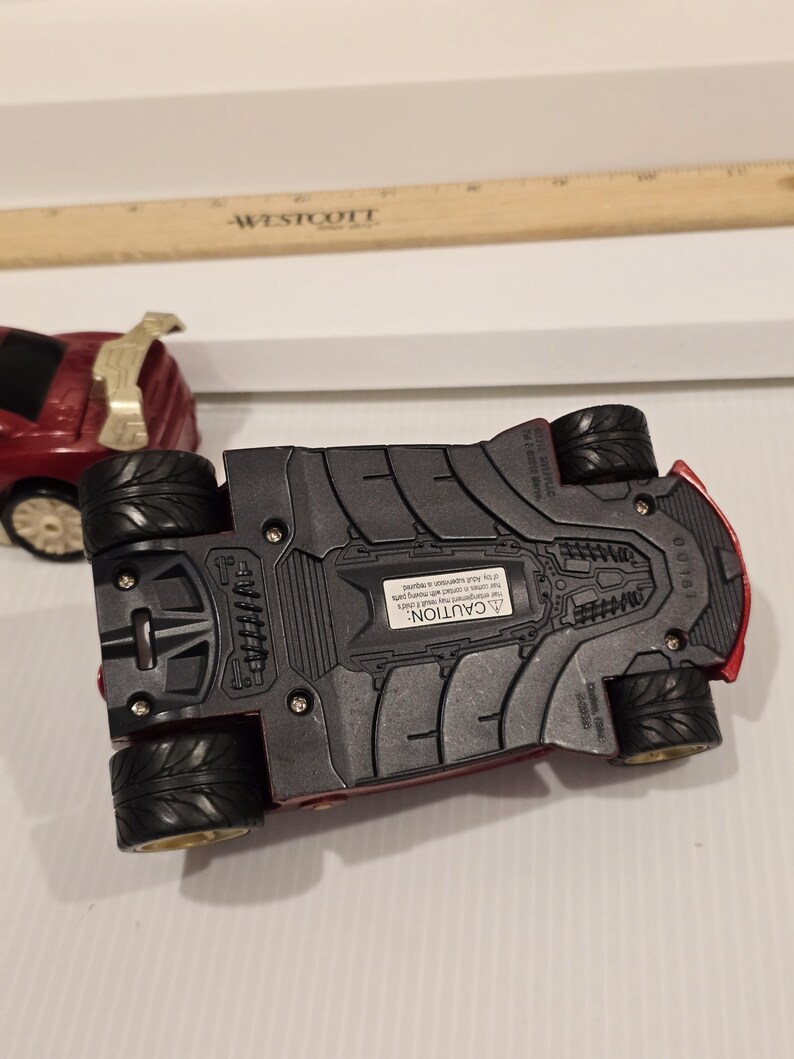 May include: A red toy car with black wheels and a gold spoiler. The underside of the car is visible, showing a textured design and a warning label. A wooden ruler is in the background.