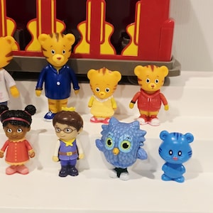 K&ouml;nnte beinhalten: Eine Sammlung farbenfroher Daniel Tiger's Neighborhood-Figuren. Die Figuren umfassen Daniel Tiger, Katerina Kittycat, O die Eule und andere Charaktere. Ein roter Trolley befindet sich im Hintergrund.
