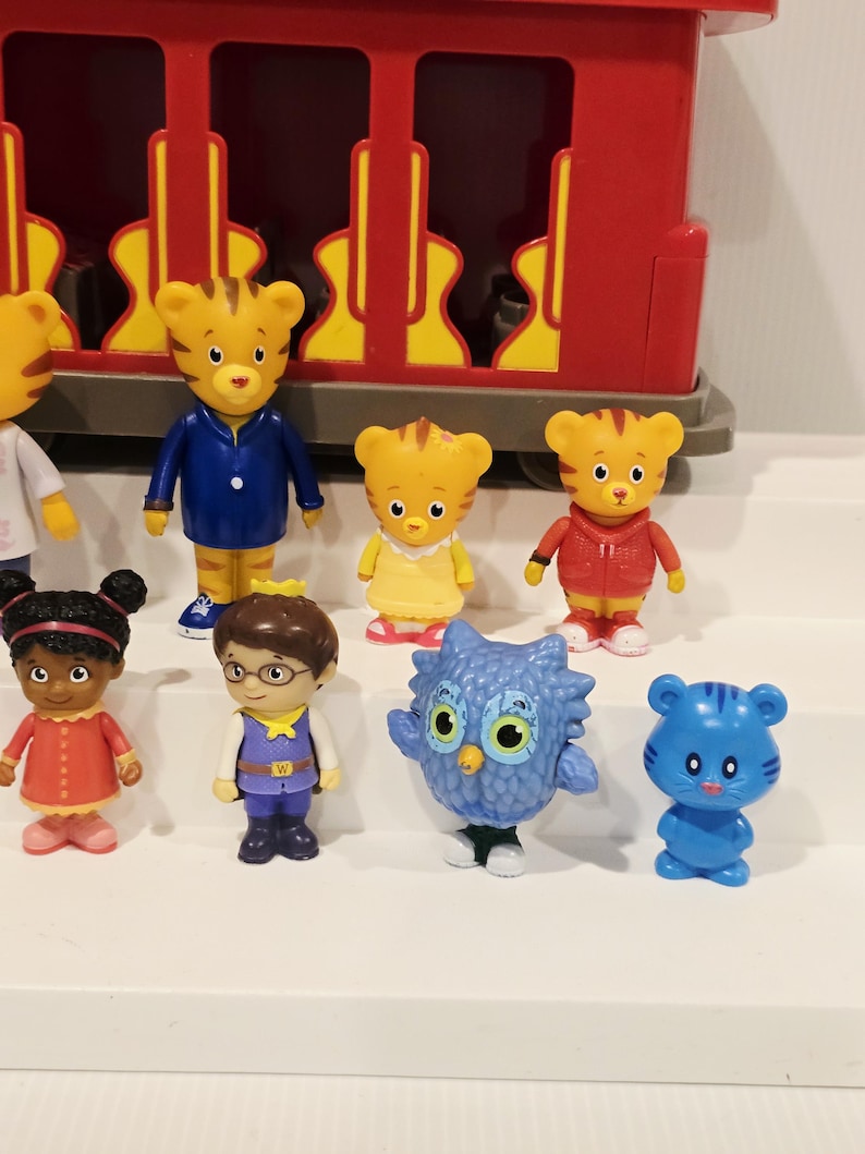 K&ouml;nnte beinhalten: Eine Sammlung farbenfroher Daniel Tiger's Neighborhood-Figuren. Die Figuren umfassen Daniel Tiger, Katerina Kittycat, O die Eule und andere Charaktere. Ein roter Trolley befindet sich im Hintergrund.