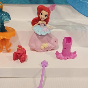 K&ouml;nnte beinhalten: Ariel-Puppe mit roten Haaren, tr&auml;gt ein rosa und blaues Kleid und ein rosa Haaraccessoire. Ebenfalls enthalten sind eine rote Krabbenfigur, eine rosa oktopusartige Figur und ein lila l&ouml;ffelf&ouml;rmiges Accessoire. Das Spielzeug befindet sich auf einer wei&szlig;en Oberfl&auml;che.