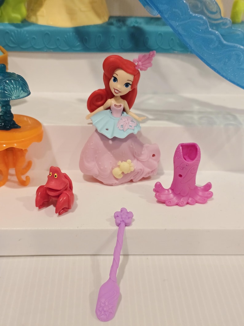 K&ouml;nnte beinhalten: Ariel-Puppe mit roten Haaren, tr&auml;gt ein rosa und blaues Kleid und ein rosa Haaraccessoire. Ebenfalls enthalten sind eine rote Krabbenfigur, eine rosa oktopusartige Figur und ein lila l&ouml;ffelf&ouml;rmiges Accessoire. Das Spielzeug befindet sich auf einer wei&szlig;en Oberfl&auml;che.