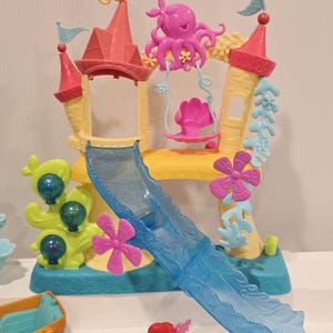 K&ouml;nnte beinhalten: Ein farbenfrohes Spielset mit einem Schloss mit Rutsche, Schaukel und verschiedenen Dekorationen zum Thema Wasser. Das Schloss hat rosa und gelbe T&uuml;rme, eine blaue Rutsche und einen rosa Oktopus. Eine kleine Ariel-Puppe ist ebenfalls enthalten.