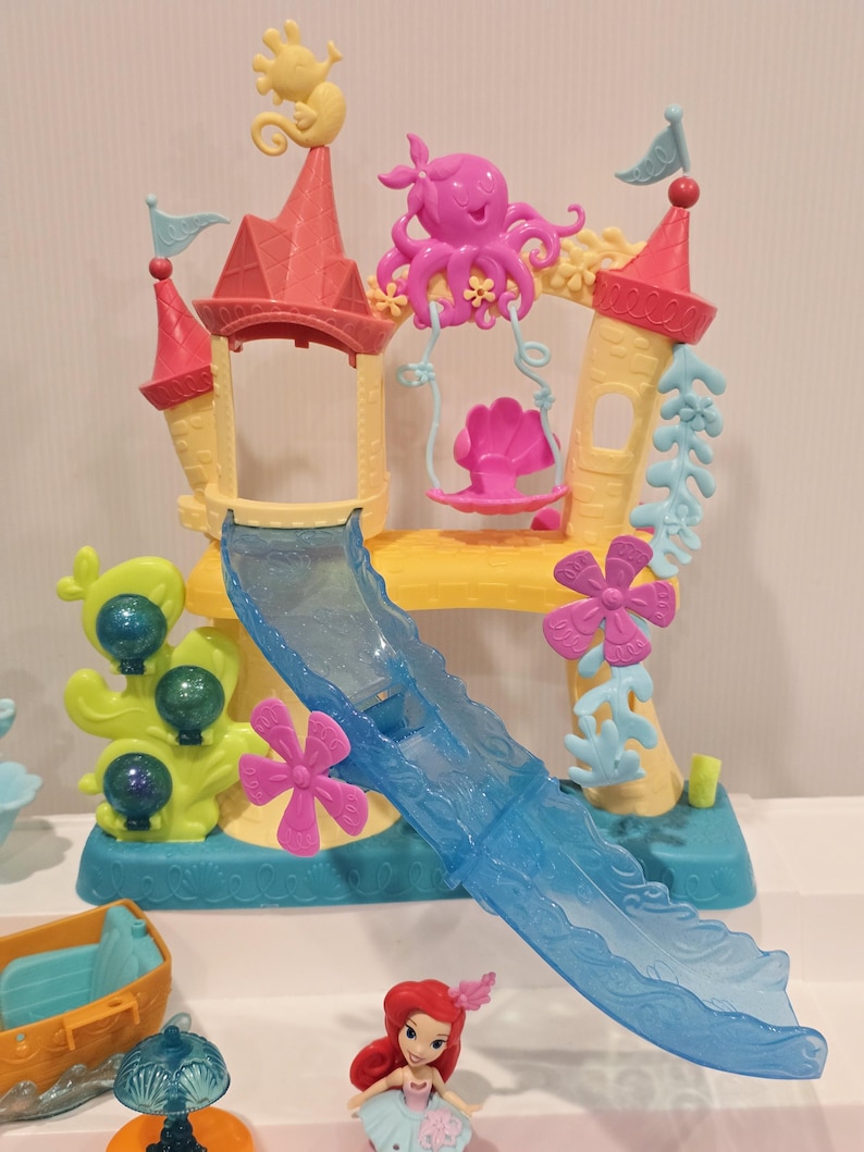 K&ouml;nnte beinhalten: Ein farbenfrohes Spielset mit einem Schloss mit Rutsche, Schaukel und verschiedenen Dekorationen zum Thema Wasser. Das Schloss hat rosa und gelbe T&uuml;rme, eine blaue Rutsche und einen rosa Oktopus. Eine kleine Ariel-Puppe ist ebenfalls enthalten.