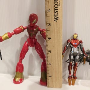 May include: Two action figures: a red Spider-Man figure with gold accents, and an Iron Man figure in red, silver, and gold. A wooden ruler is positioned between them, measuring approximately 6 inches.