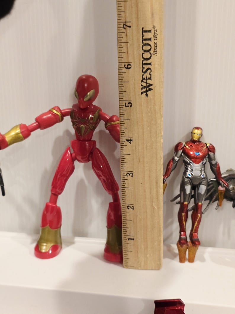 May include: Two action figures: a red Spider-Man figure with gold accents, and an Iron Man figure in red, silver, and gold. A wooden ruler is positioned between them, measuring approximately 6 inches.