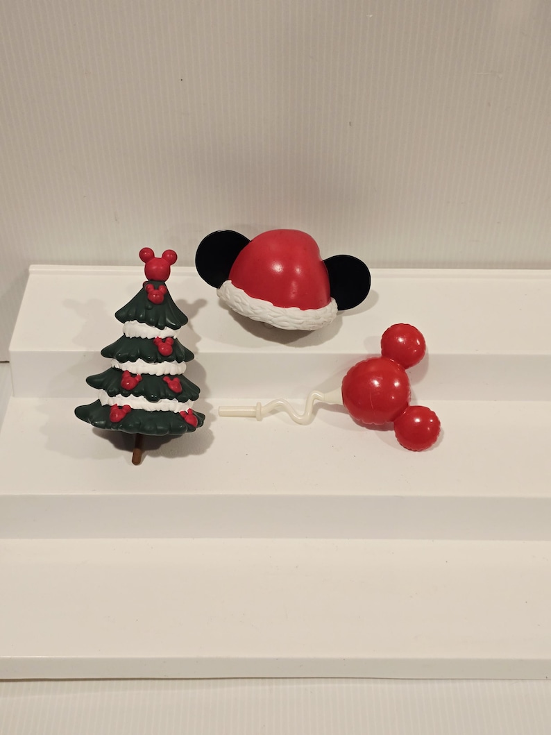 May include: A set of three small plastic ornaments including a dark green Christmas tree decorated with red mouse-shaped icons, a red Santa hat with black mouse ears, and a red mouse-shaped balloon on a thin white stick with a wavy stem.