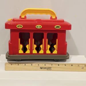 K&ouml;nnte beinhalten: Ein roter Spielzeugzug mit gelbem Griff und Akzenten. Das Spielzeug hat drei &Ouml;ffnungen mit gelben und schwarzen Designs. Ein Holzlineal ist vor dem Spielzeug platziert. Der Spielzeugzug ist etwa 30 cm lang.