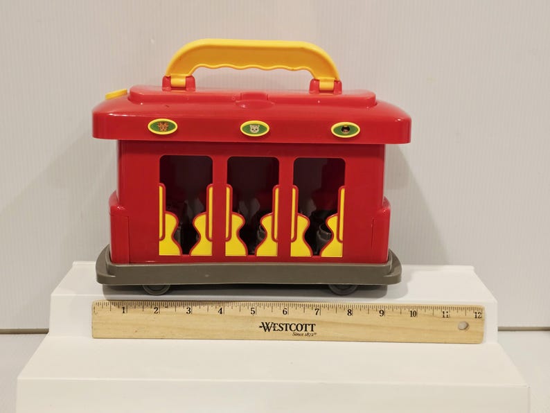K&ouml;nnte beinhalten: Ein roter Spielzeugzug mit gelbem Griff und Akzenten. Das Spielzeug hat drei &Ouml;ffnungen mit gelben und schwarzen Designs. Ein Holzlineal ist vor dem Spielzeug platziert. Der Spielzeugzug ist etwa 30 cm lang.