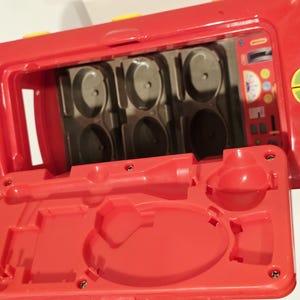 K&ouml;nnte beinhalten: Ein rotes Kunststoffspielzeug mit offenem Deckel, das F&auml;cher und geformte Formen freigibt. Das Spielzeug hat gelbe Akzente und einen detaillierten Innenraum mit verschiedenen geformten Merkmalen. Der offene Deckel ist ebenfalls rot.