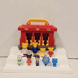 K&ouml;nnte beinhalten: Ein roter Spielzeugzug mit gelbem Griff und mehrere kleine Figuren. Die Figuren umfassen einen Tiger, eine Eule und andere Tierfiguren in verschiedenen Outfits. Das Spielzeug ist auf einer wei&szlig;en, abgestuften Plattform angeordnet.