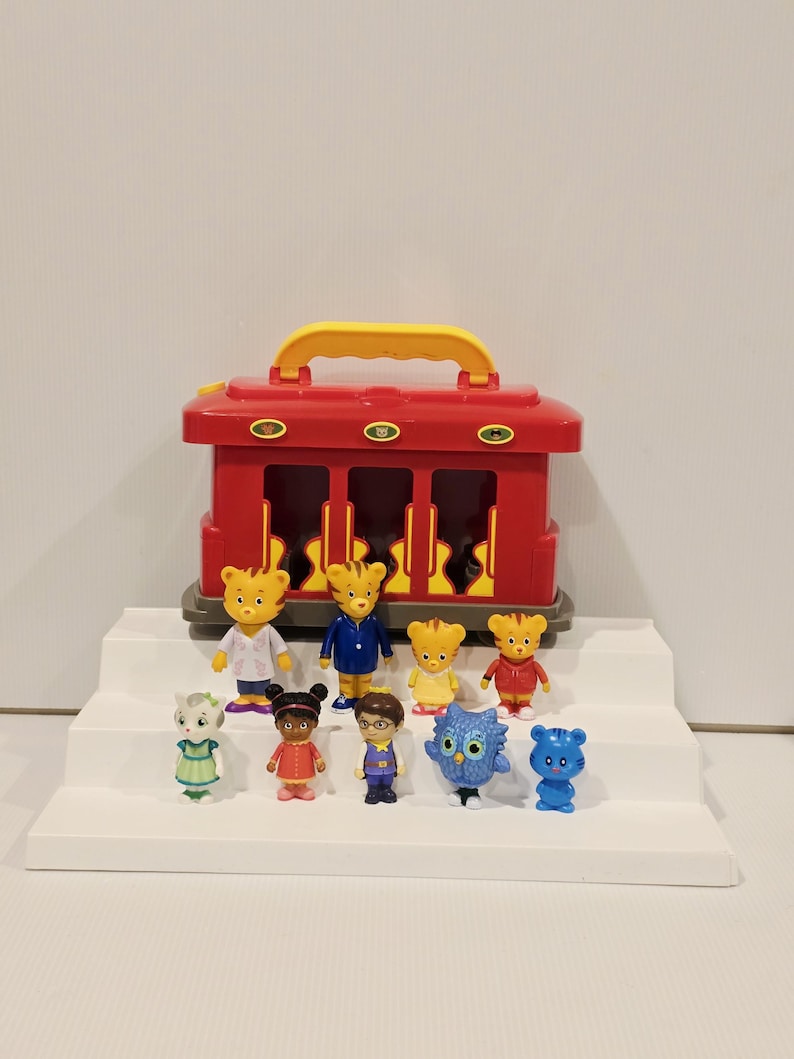 K&ouml;nnte beinhalten: Ein roter Spielzeugzug mit gelbem Griff und mehrere kleine Figuren. Die Figuren umfassen einen Tiger, eine Eule und andere Tierfiguren in verschiedenen Outfits. Das Spielzeug ist auf einer wei&szlig;en, abgestuften Plattform angeordnet.